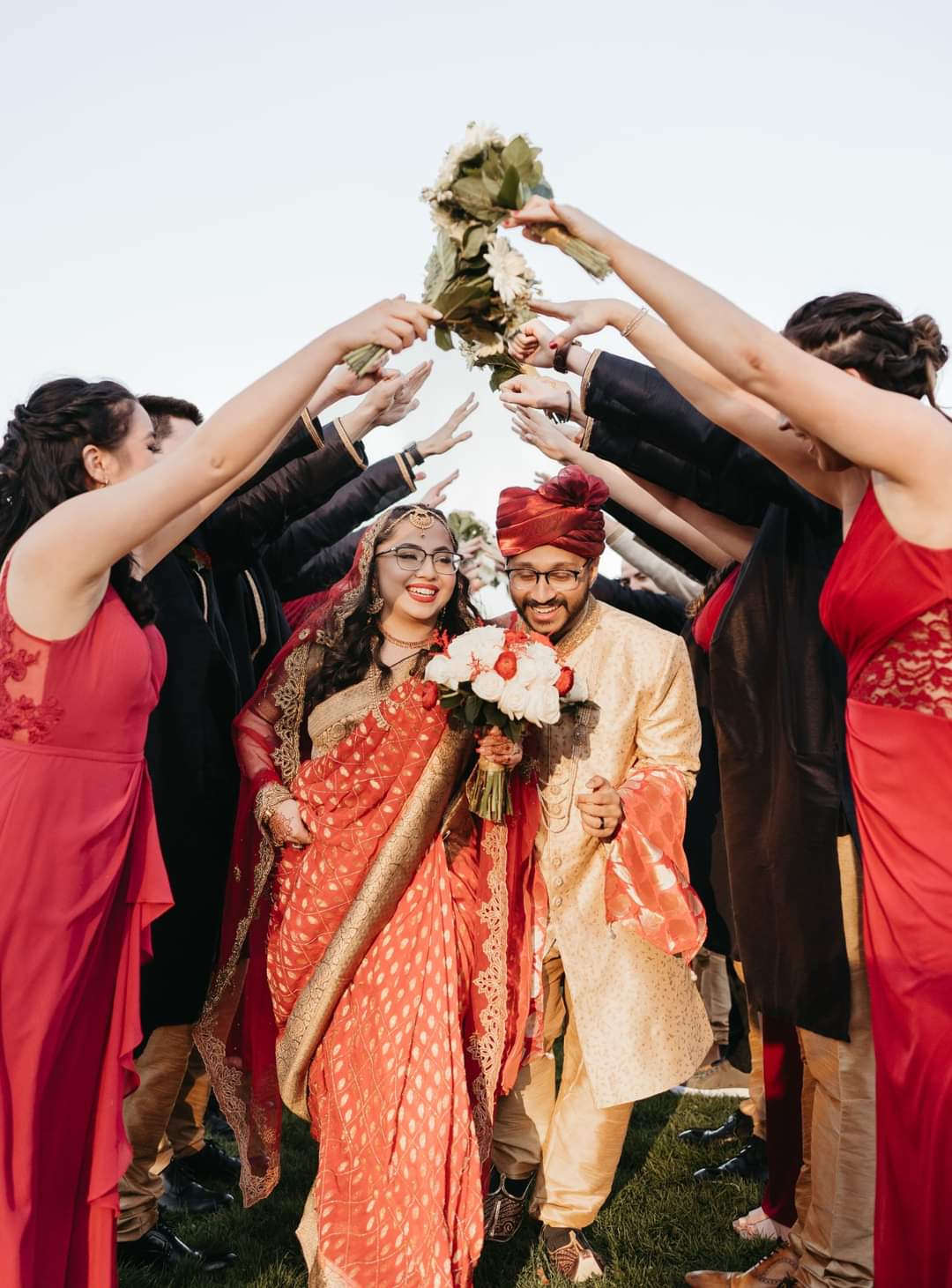 Indian bride in a red and gold saree and groom in a sherwani and turban walk through a cheering bridal party at a multicultural Boston wedding with DJ Angel Alba